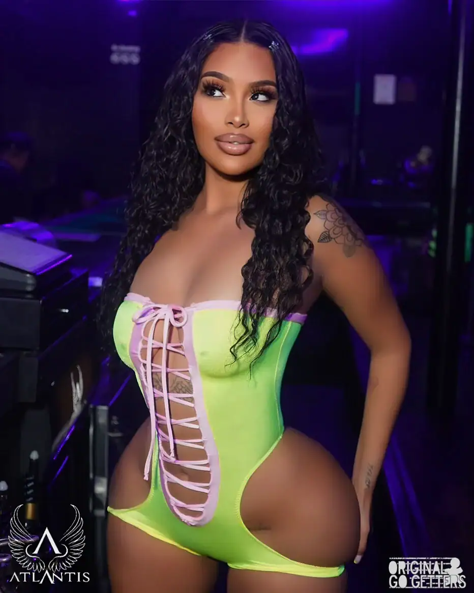 A woman with long curly hair poses in a neon green and pink lace-up bodysuit in a dimly lit nightclub, capturing the vibrant energy of adult entertainment Jamaica, NY. Logos for "Atlantis" and "Original Go Getters" appear in the corners.