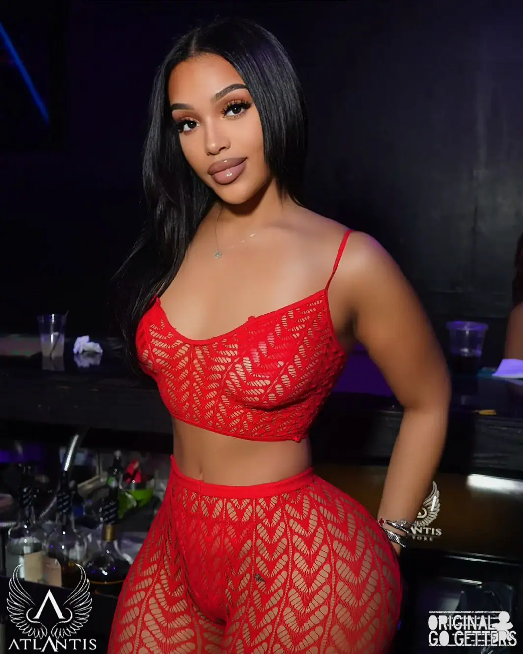 A woman with long, straight black hair poses at a bar in a strip club Jamaica, wearing a matching red lace crop top and high-waisted pants. The nightclub setting features bottles on the counter and club logos in the corners.
