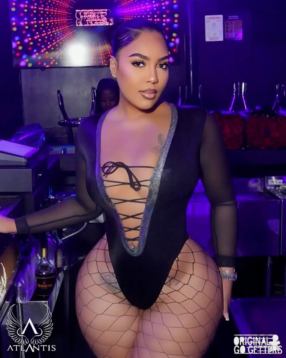 A woman poses confidently in a nightclub, wearing a deep V-neck black bodysuit with lace-up details and fishnet stockings, capturing the vibrant vibe of adult entertainment Jamaica. Colorful lights and club decor set the scene; Atlantis and Original GoGetters logos appear.
