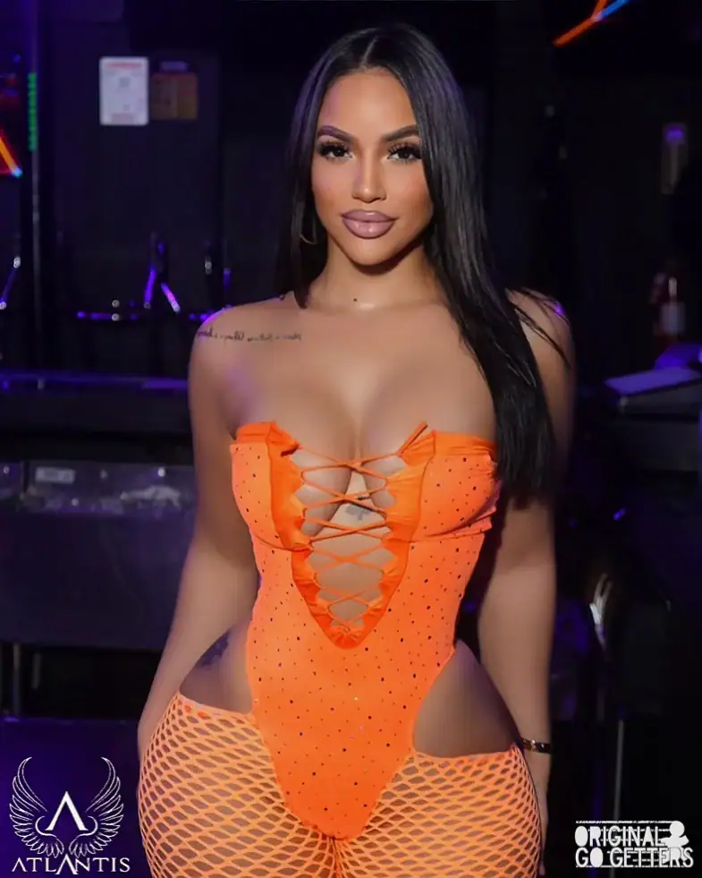 A woman with long black hair wears a bright orange, lace-up bodysuit and matching fishnet stockings in a dimly lit strip club Jamaica, NY. Logos for Atlantis and Original Go Getters are visible in the corners.