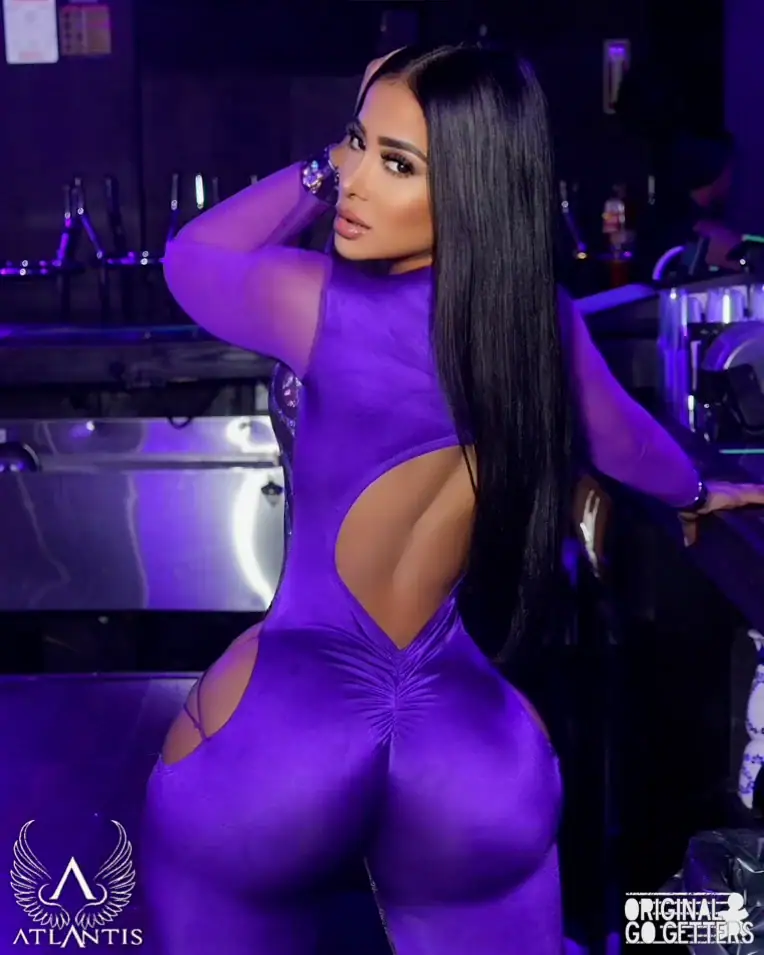 A woman with long straight hair wears a vibrant purple, backless bodysuit, posing with her back to the camera in a lively nightclub setting. The Atlantis logo appears in the lower left corner, capturing the allure of adult entertainment Jamaica nightlife.