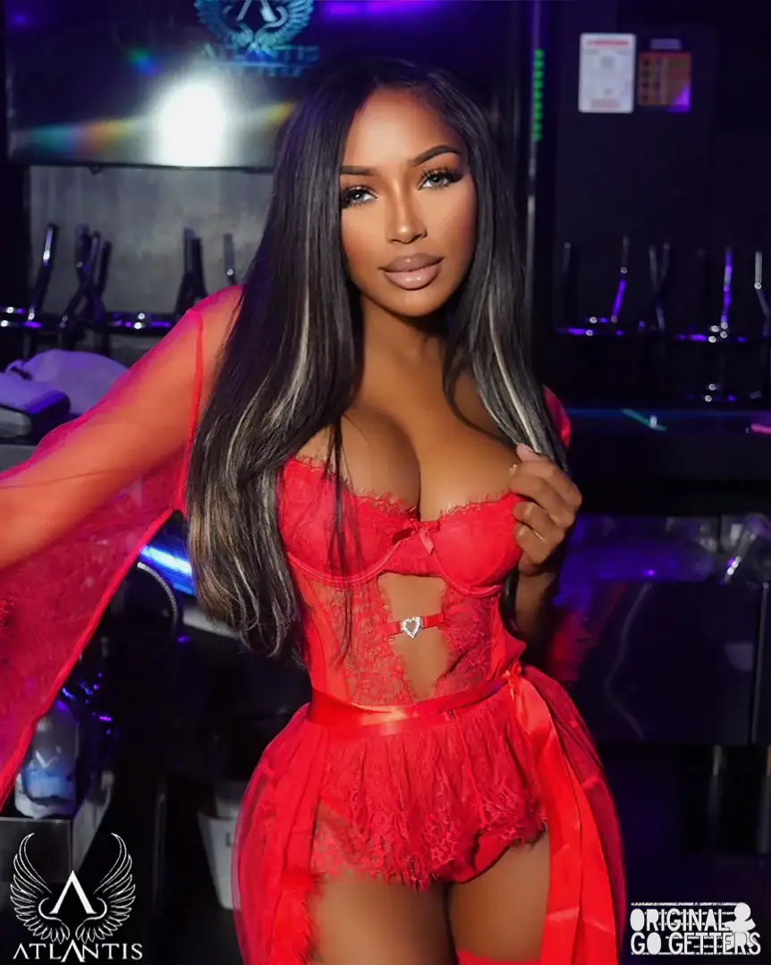 A woman with long straight hair wears a sheer red lace lingerie set and robe, standing in a nightclub with a dark, neon-lit background—capturing the allure of adult entertainment Jamaica. Logos for “Atlantis” and “Original Go Getters” appear at the bottom corners.